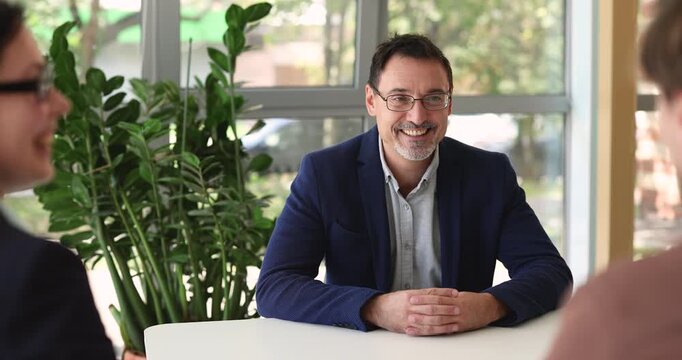 Middle-aged man sitting at table in front of two HR managers, answers questions about professional skills and experience during job interview in office. Company staffing, human resources, employment