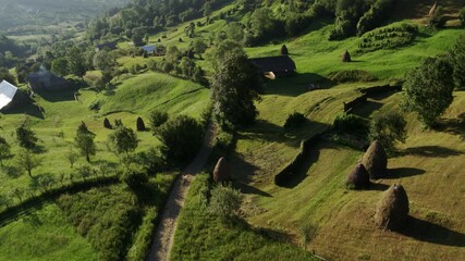 Scenic drone shot of a peaceful countryside landscape featuring rolling green hills, a dirt path, and haystacks.