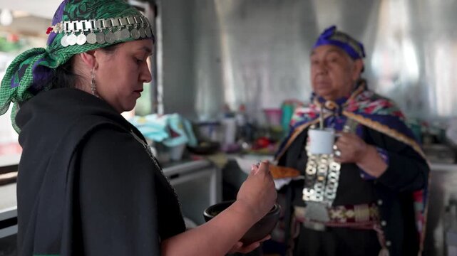 Two native mapuche women in traditional clothing sharing a meal inside a local kitchen