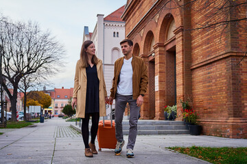 Man and woman walking together with suitcase near brick building