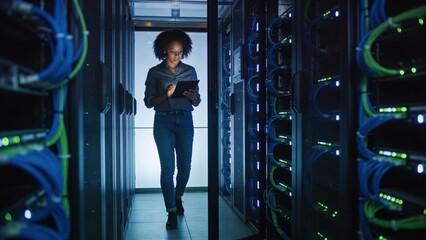 A woman in business attire walks confidently down a server room hallway, looking at a tablet. It symbolizes technological advancement, data management, and a modern, digital workplace.