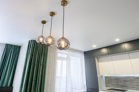 modern kitchen with sleek grey cabinetry, grey brick-style backsplash, three spherical pendant lights with brass fixtures, emerald velvet curtains, and sheer white drapes