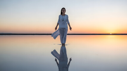 A woman standing in white clothes on a calm water surface reflecting the sunset sky in a meditative pose