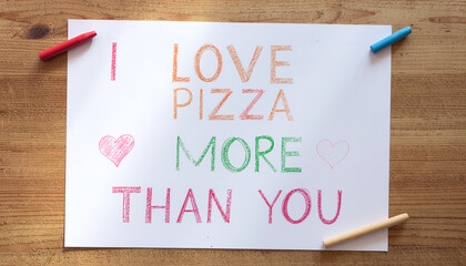 "I Love Pizza More Than You" funny hand-drawn quote in colorful crayon on white paper with wax crayons on wooden table background for National Pizza Day.