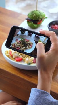 Food blogger taking a photo of pasta and a green burger with a mobile phone camera for her social media. Trendy culinary vlogging and content creation at a restaurant with healthy gourmet meal