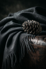 A meticulously detailed shot showcases a textured, dark gray scarf with fringe, artfully draped over a vintage leather ottoman, alongside a single pinecone.