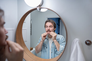Young man with mustache looks at himself in mirror before work, morning self-care routine. Self-confidence male standing in bathroom checking face skin condition. Modern masculinity, self improvement.