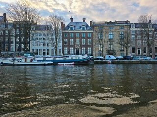 Winter view of an Amsterdam canal with historic houses, snow-covered houseboats, bare trees, and parked cars, capturing a quiet, tranquil European cityscape scene