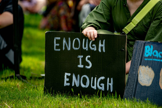 A protester in green clothing holds a black sign with white text reading "Enough is Enough" during an outdoor rally or demonstration on grass with other protesters in background.