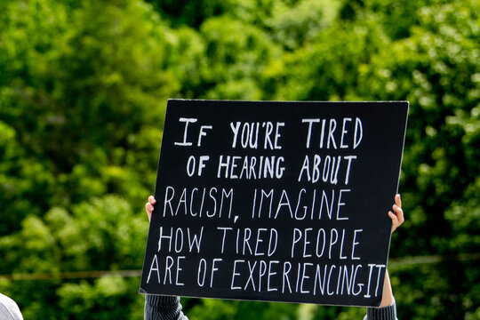 Person holding black protest sign with white text message about experiencing racism at outdoor demonstration . Shows activism and social justice messaging at peaceful protest.