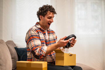 Happy young man smiling and opening delivered package with headphones while sitting on sofa at his home. Image captures joy and convenience of home shopping and receiving order.	