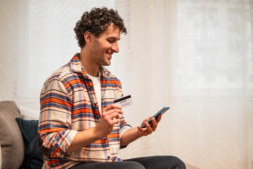 Smiling young man sitting on sofa in living room, comfortably using his smartphone for mobile payment with credit card. Wireless technology and online shopping concept.