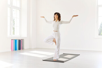 Yoga balance asana pose with mindfulness. In a sunlit studio, a woman holds a tree variation, arms wide, holding stability while focusing on breath. Balance, calm focus, and inner harmony.