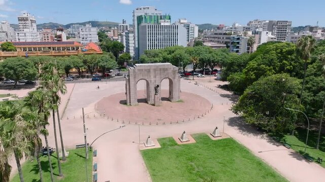 Cinematic aerial drone flyover revealing the Monumento ao Expedicionario arch behind palm trees in Farroupilha Park. Historic landmark in Porto Alegre. D-Log 10-bit 4K video.