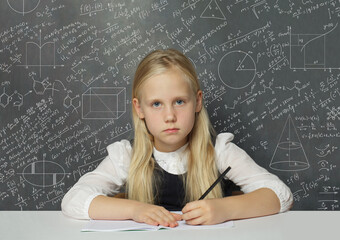 Unhappy child student sitting in classroom on chalkboard background with mathematical formulas