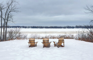 Three snow covered Adirondack chairs sitting in the winter snow by the shores of the Ottawa river in Canada