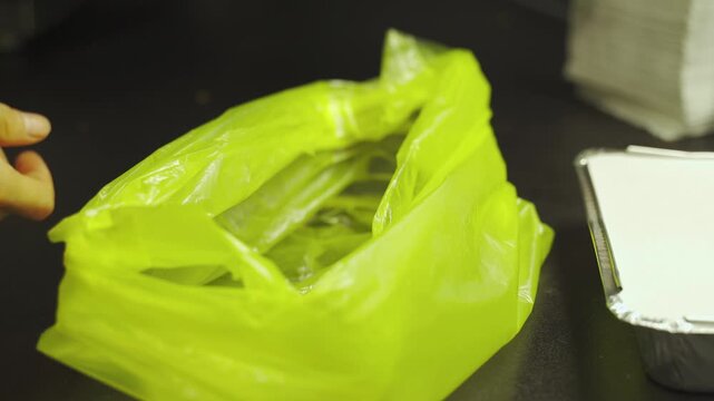 Hands skillfully pack hot cheburek into a shiny foil box, sealing in warmth and flavor. A vibrant green bag awaits to hold the tasty treat, ready for that perfect meal.