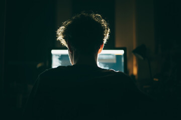 A young man is seated at a computer in a dark room, illuminated solely by the screen light, creating a minimal and modern tech atmosphere.
