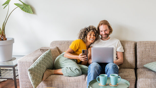 Multiracial young couple watching computer laptop sitting on the sofa at home - Happy diverse husband and wife using pc online services - Technology life style concept