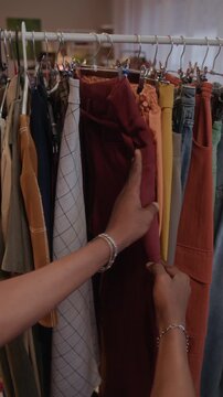 Vertical POV shot of unrecognizable shopper examining authentic items hanging on clothing rack in modern secondhand shop with warm lighting and cozy atmosphere