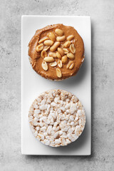 Puffed rice cakes with peanut butter and peanuts on a white plate, grey background. Top view