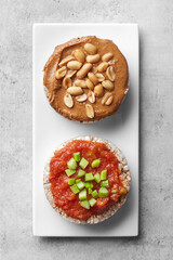 Puffed rice cakes with tomato paste, celery, peanut butter and peanuts on a white plate, grey background. Top view