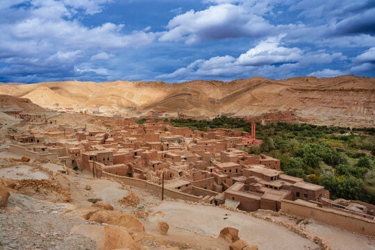 Ancient desert village with adobe houses and lush greenery under a dramatic cloudy sky. Haut Atlas, Ounila Valley, Morocco