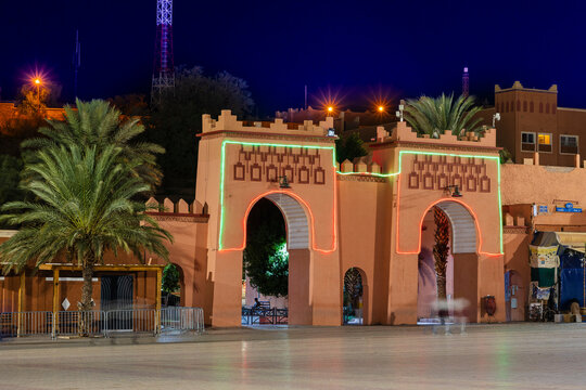 Brightly lit historic gate with neon lights framed by palm trees at night under deep blue sky. Ouarzazate, Morocco