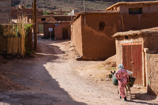 A person carrying items in a village street with rustic, earthen buildings in bright daylight. Ouarzazate, Morocco