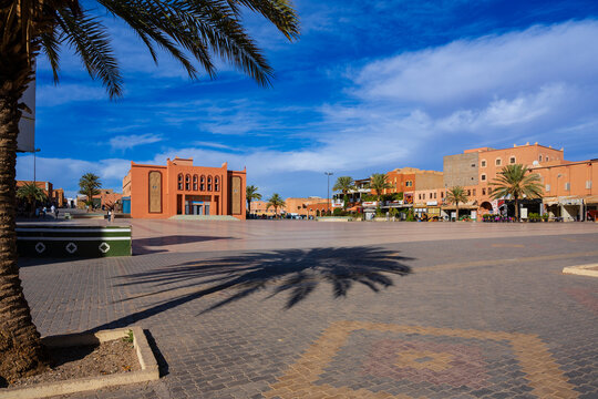 A sunny plaza with palm trees casts shadows on a patterned brick courtyard against a bright blue sky. Ouarzazate, Morocco