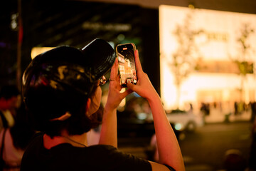 Woman captures city lights at night with a smartphone, wearing a black cap for style. Chengdu, Sichuan, China