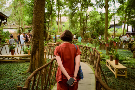 A person in red clothing walks down a tree-lined path in a lush garden. Chengdu, Sichuan, China - Powered by Adobe
