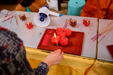 A person lights candles on a tray at a colorful indoor ritual setup with incense on tables. Chengdu, Sichuan, China