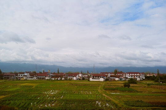Wide view of lush green fields and a distant village beneath a cloudy blue sky. Chengdu, Sichuan, China