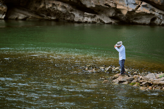Person fishing on rocky riverbank with forested cliff in background. Li River, Li Jiang, Guangxi, China