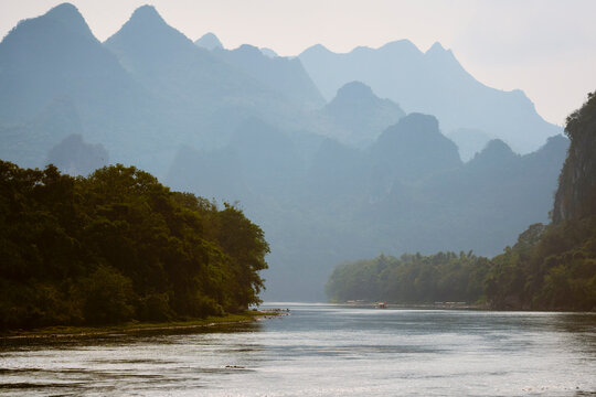 Serene river scene framed by lush trees and distant mountain silhouettes under a soft sky. Li River, Li Jiang, Guangxi, China