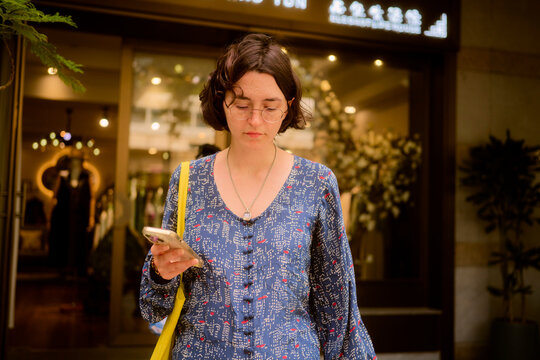Woman with glasses and blue dress looking at phone outside a store with bright lighting. Guangzhou, Guangdong, China