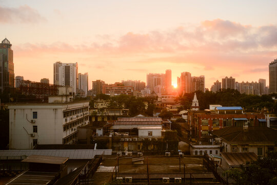 A vibrant city skyline at sunset with sun peeking through high-rises and scattered clouds. Guangzhou, Guangdong, China