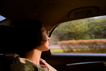 Person with glasses gazing out a car window during a rainy day with blurred greenery outside. Trip from Guilin to Chengdu, Guangxi, Sichuan, China