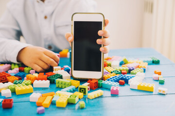 Fototapeta premium Child hand showing mobile phone with blank white screen with colorful toy bricks