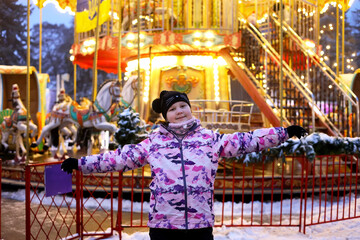 Happy girl in pink jacket posing near illuminated carousel, child enjoying winter evening at bright amusement park fairground during holidays