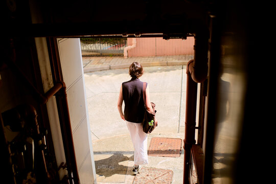 A person in a sleeveless top and white pants exits a building onto a sunlit street. Hongkong, China