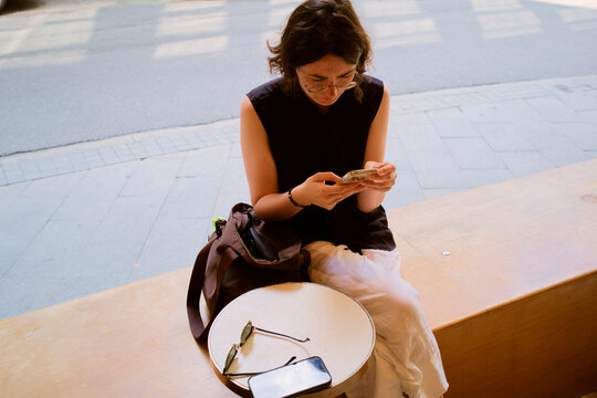 Woman sits on a bench by the road, focused on counting cash at a table with a phone and glasses. Hongkong, China