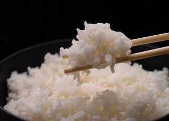 Close up of chopsticks holding steamed white rice over a bowl on black background