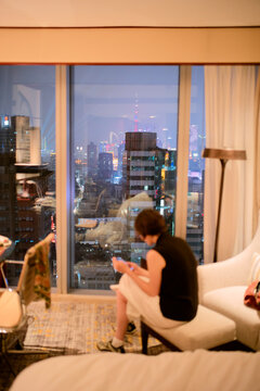 Woman sitting by a hotel room window overlooking a cityscape at night, reading a book.   Shanghai, China