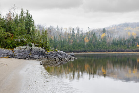 Tranquil lakeside with rocky shore, pine forest, and autumn foliage under cloudy skies. Thunder Bay, Ontario, Canada