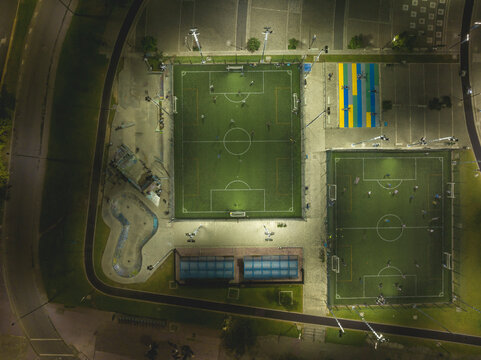 Aerial view of illuminated outdoor soccer fields at night with players and surrounding parking. Bogota DC, Colombia