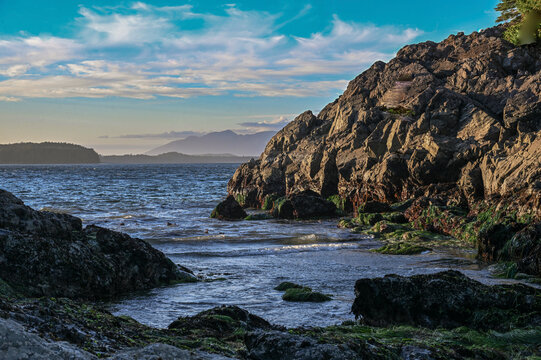 Rocky coastline with blue skies, distant mountains, and gently lapping ocean waves. Tofino, British Columbia, Canada