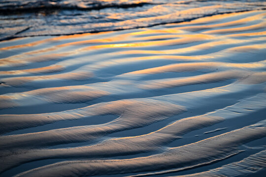 Ripples in wet sand reflect the soft glow of a sunset over the ocean waves. Tofino, British Columbia, Canada
