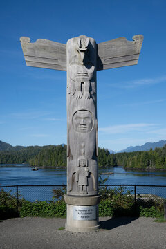 Wooden totem pole stands by a lake with mountains and trees in the background under clear sky. Tofino, British Columbia, Canada